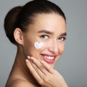 Beautiful woman with cream in heart shape on her cheeks representing skin care concept